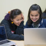 Two freshmen students hudding around a laptop screen laughing.